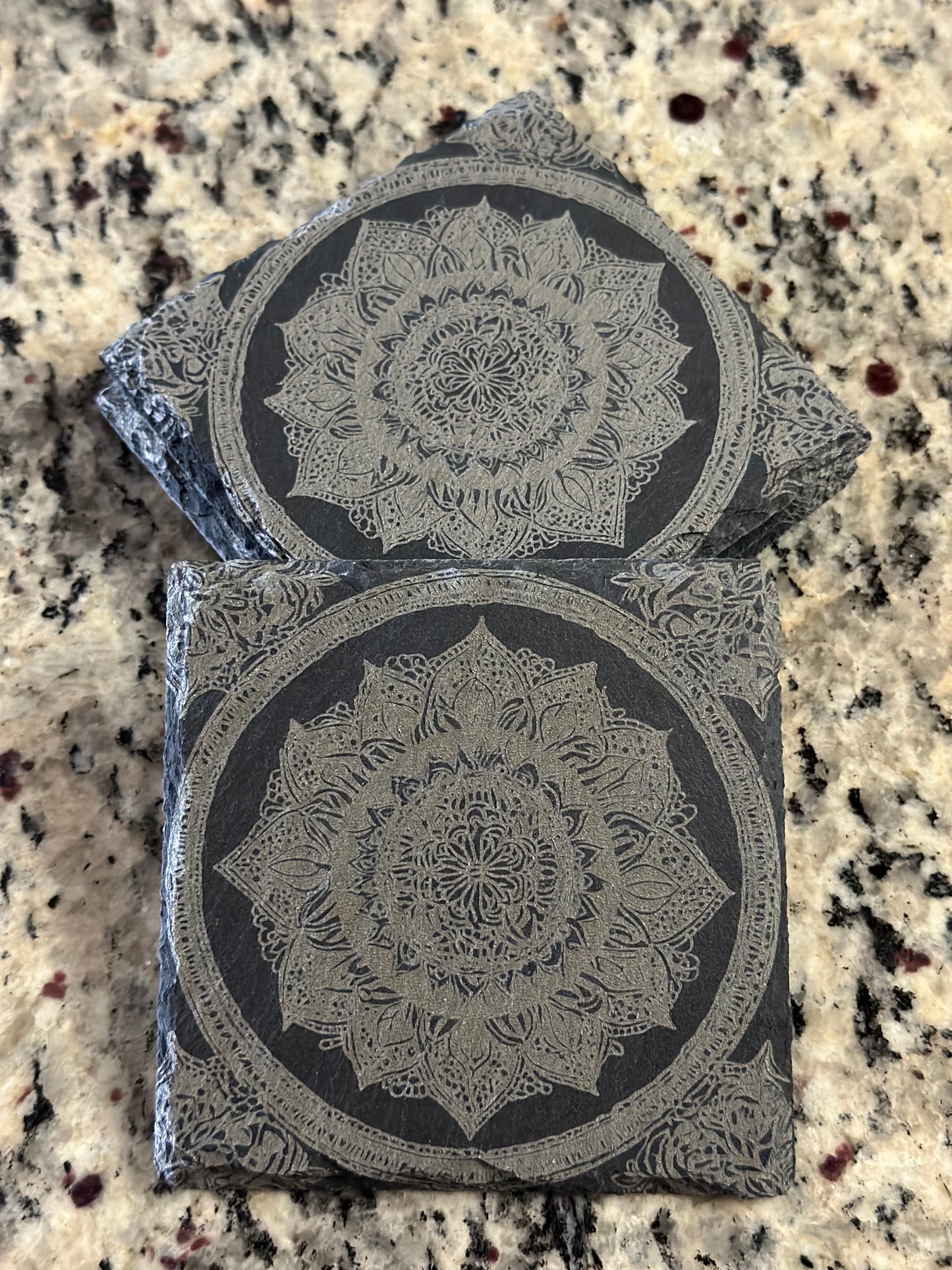 Engraved slate coasters with detailed mandala style image on a dark slate background.