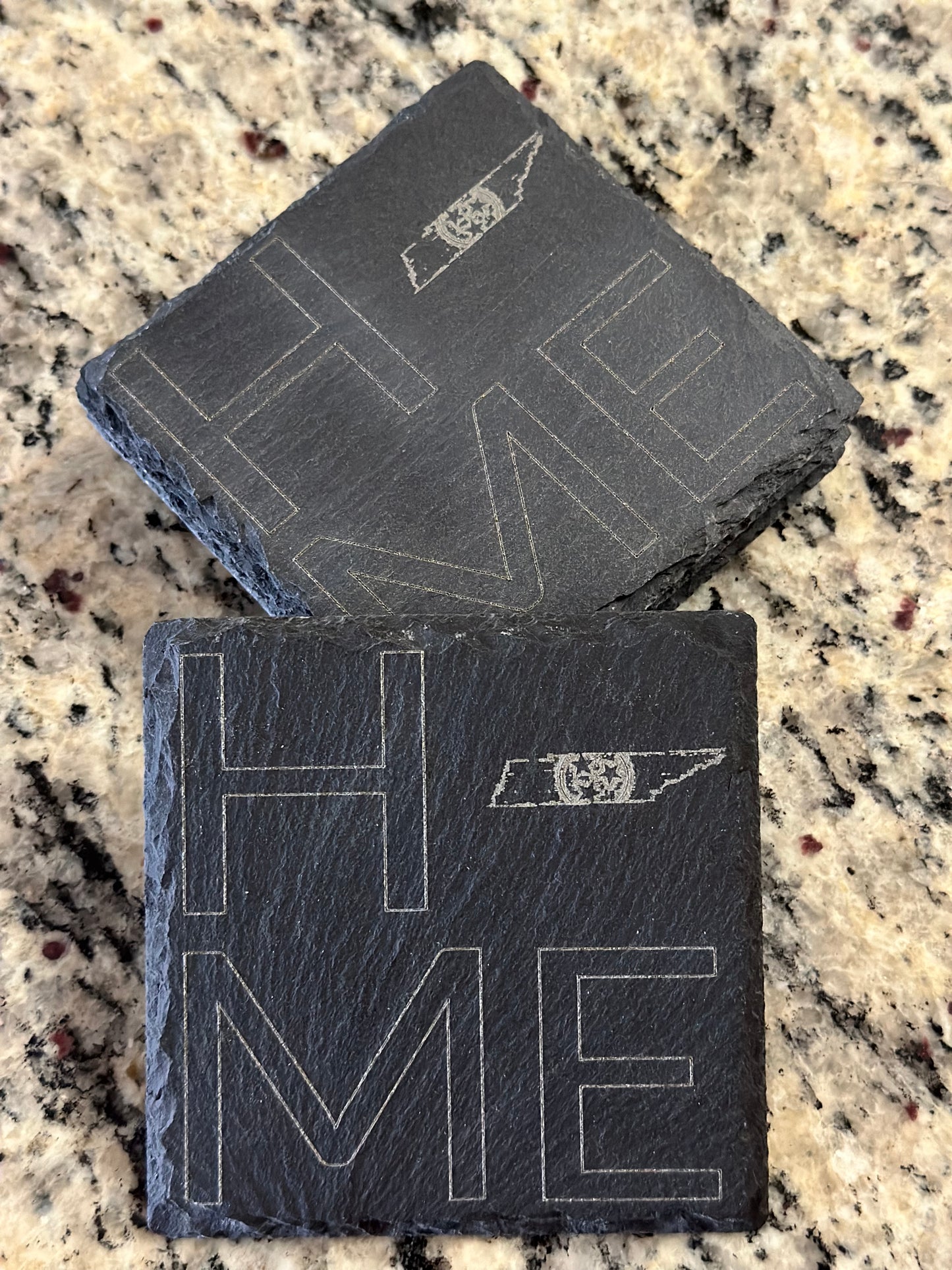 Engraved slate coasters with 'HOME' with TN State LOGO in place of O text on a dark slate background.