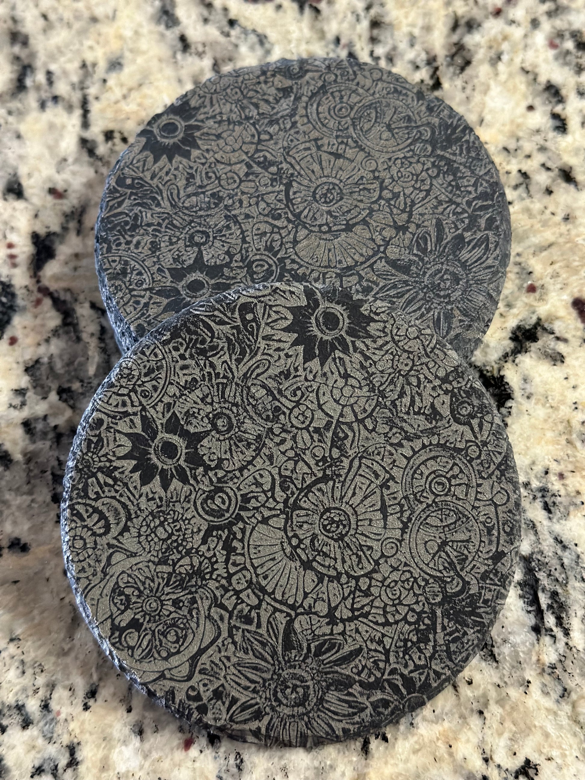 A black slate coaster with an engraved pattern of Steampunk Flowers.